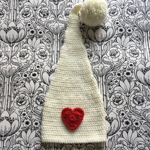 Pode incluir: Chapéu de gnomo de crochê branco com um coração vermelho e um pompom branco no topo.