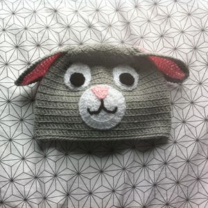 Könnte beinhalten: Graue Häkelmütze mit rosa Ohren und einem weißen Häkelgesicht mit schwarzen Augen und einer rosa Nase.