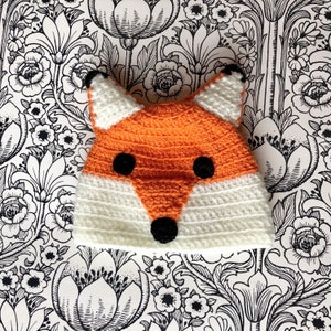 Könnte beinhalten: Eine gehäkelte Fuchs-Mütze aus orangefarbenem und weißem Garn. Die Mütze hat schwarze Filzaugen und eine schwarze Filznase.