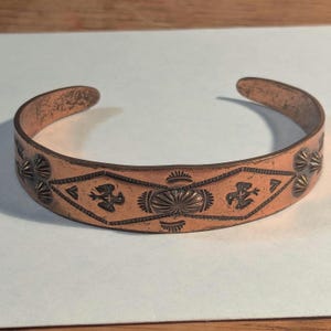 May include: A copper cuff bracelet featuring a central oval stamp surrounded by geometric lines and bird motifs. The metal surface displays a warm, reddish-brown tone with dark, recessed stamped details throughout the band's exterior.