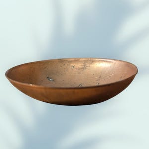 Puede incluir: Un cuenco circular poco profundo con un acabado de cobre metálico. La superficie interior muestra un aspecto desgastado y texturizado con sutiles manchas oscuras y finos arañazos en la base, con un tono marrón rojizo uniforme.