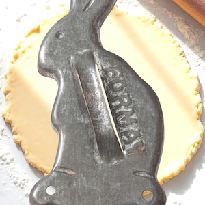 May include: A vintage metal bunny-shaped cookie cutter with the word "FORM" stamped on the side. The cutter is gray and sits on a light-colored surface with a rolled-out piece of dough and a wooden rolling pin.