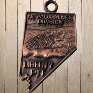 May include: A copper-colored metal pendant in the shape of Nevada, featuring the text "NEVADA MINES DIVISION" and "LIBERTY PIT." The pendant has a detailed relief of a landscape and a circular loop at the top for hanging.