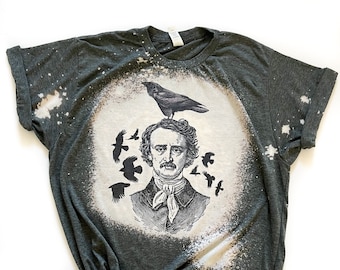 Op de afbeelding: Een donkergroen T-shirt met een ontwerp van bleekmiddelspatten met een zwart-witte illustratie van Edgar Allan Poe met een raaf op zijn hoofd. Rondom Poe bevinden zich verschillende kleinere raven.