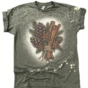 Può includere: Una t-shirt a maniche corte verde oliva scuro con un effetto a spruzzi candeggiati. La grafica centrale presenta un'illustrazione dettagliata di pigne, rametti di pino, anice stellato e bastoncini di cannella legati con dello spago.