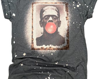 Puede incluir: Una camiseta gris oscuro de manga corta con efecto desteñido y un estampado gráfico rectangular en el pecho. El estampado muestra un retrato en blanco y negro del monstruo de Frankenstein soplando una burbuja de chicle grande y rosa.