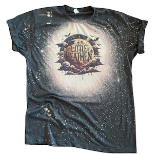 Peut inclure: Un t-shirt à manches courtes gris anthracite foncé avec un effet délavé, présentant un motif central composé d'une grande lune, de nuages et de chauves-souris. Le texte "LITTLE HEATHEN" est imprimé au centre dans une police stylisée.