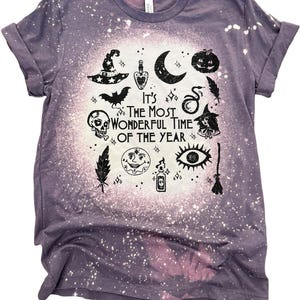 May include: A purple short-sleeve t-shirt with a bleached center effect. The graphic features black illustrations of a witch hat, moon, pumpkin, skull, bat, snake, feather, sun, eye, and broom, with the text IT'S THE MOST WONDERFUL TIME OF THE YEAR.