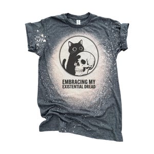 May include: A dark heather gray short-sleeve t-shirt with a bleached circular pattern in the center. The graphic features a black cat sitting next to a human skull inside a circle, with the text EMBRACING MY EXISTENTIAL DREAD printed below.