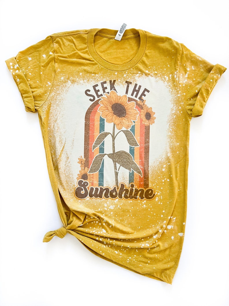 May include: A mustard yellow t-shirt with a bleached effect. The shirt features a graphic of a sunflower with a rainbow behind it and the text "SEEK THE SUNSHINE".