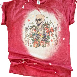 May include: Red t-shirt with a bleached white circle design. The graphic features a skeleton wearing Christmas lights, surrounded by gifts and holly. The skeleton is making a rock and roll hand gesture.