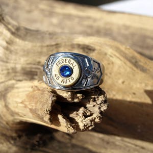 Könnte beinhalten: Ein silberner Ring mit goldfarbenem Mittelteil, der den Text "FEDERAL 45 AUTO" und einen blauen Edelstein trägt. Der Ring ist vor einem natürlichen Holzuntergrund platziert und zeigt sein aufwendiges Design.