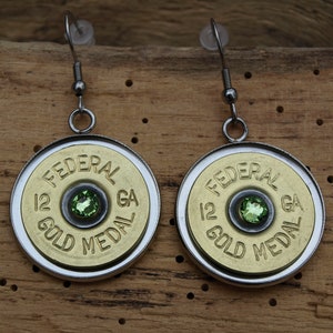 Könnte beinhalten: Ein Paar silberne Ohrringe mit einem goldfarbenen Schrotflintenpatronen-Anhänger mit dem Text "FEDERAL 12 GA GOLD MEDAL" und einem grünen Edelstein in der Mitte.