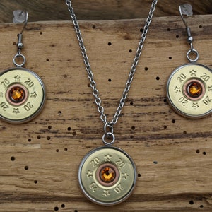 Puede incluir: Un conjunto de joyas de casquillo de bala de color dorado con un strass naranja claro en el centro de cada pieza. El conjunto incluye un collar y un par de pendientes.