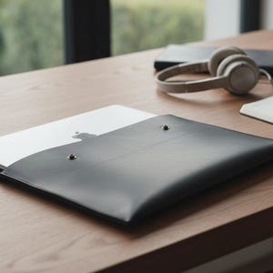 Könnte beinhalten: Eine schwarze Laptop-Hülle aus Leder mit zwei silbernen Druckknöpfen. Ein weißer Laptop ist teilweise in der Hülle sichtbar. Weitere Gegenstände auf dem Holztisch sind Kopfhörer, ein Notizbuch und ein Stift.