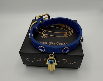 May include: A blue leather dog collar with gold accents and a hamsa hand charm with a blue evil eye. The collar has blue evil eye studs around the edge. The collar is on a black box with the text "Crystal Pet Collar".