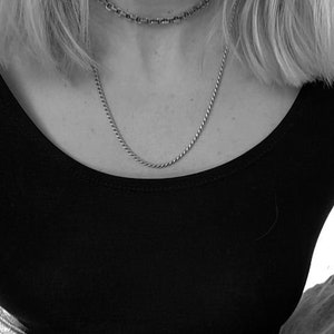 May include: A close-up of a person wearing a black scoop neck shirt and two silver necklaces. The person is wearing blue jeans.