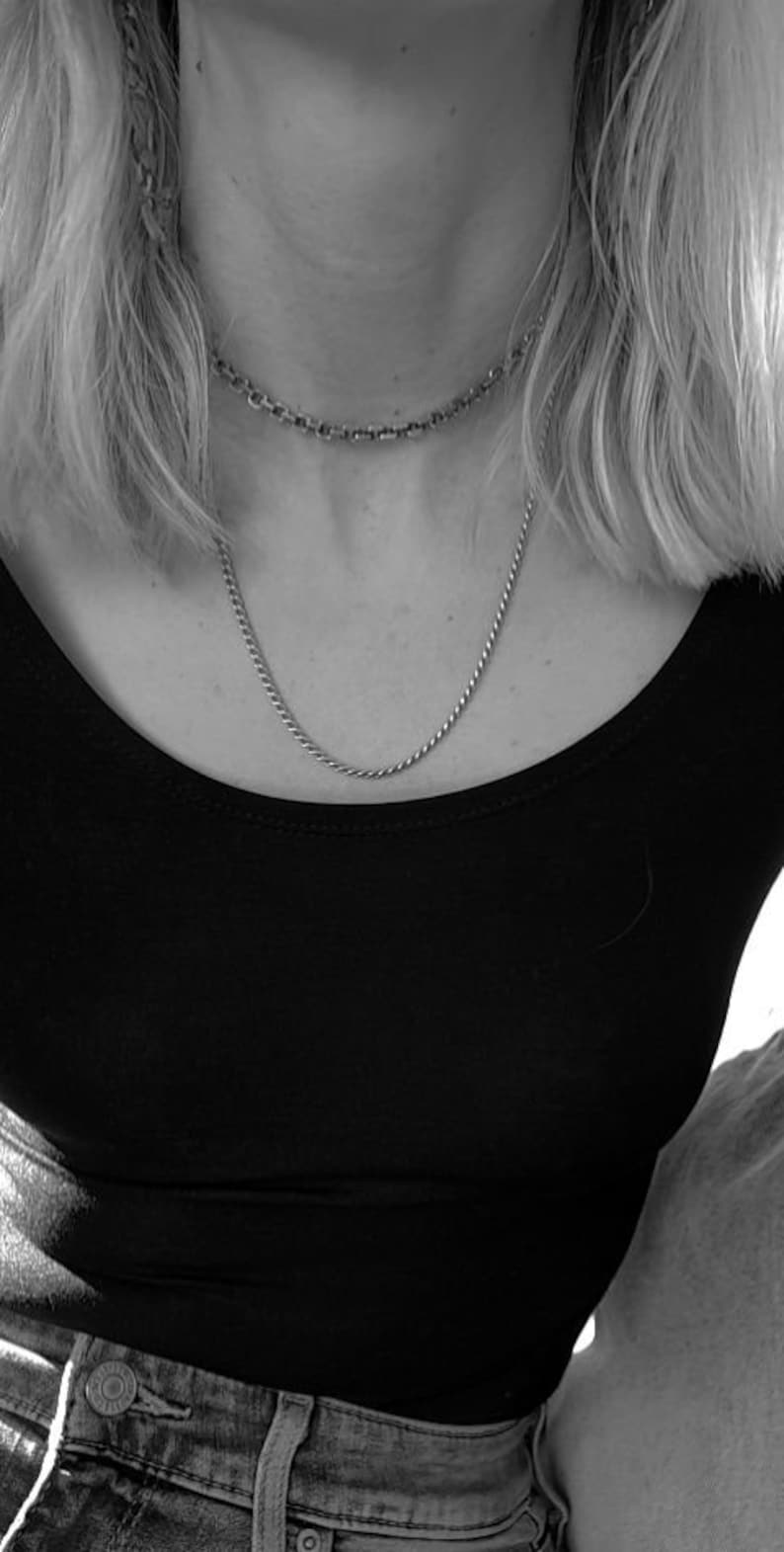 May include: A close-up of a person wearing a black scoop neck shirt and two silver necklaces. The person is wearing blue jeans.