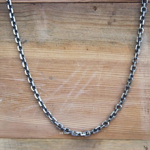 May include: A silver chain necklace with a box chain design. The chain is laying on a wooden surface.