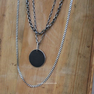 May include: Three silver chains with a round, black pendant. The chains are layered on top of each other and the pendant is hanging from the bottom chain. The chains are on a wooden surface.