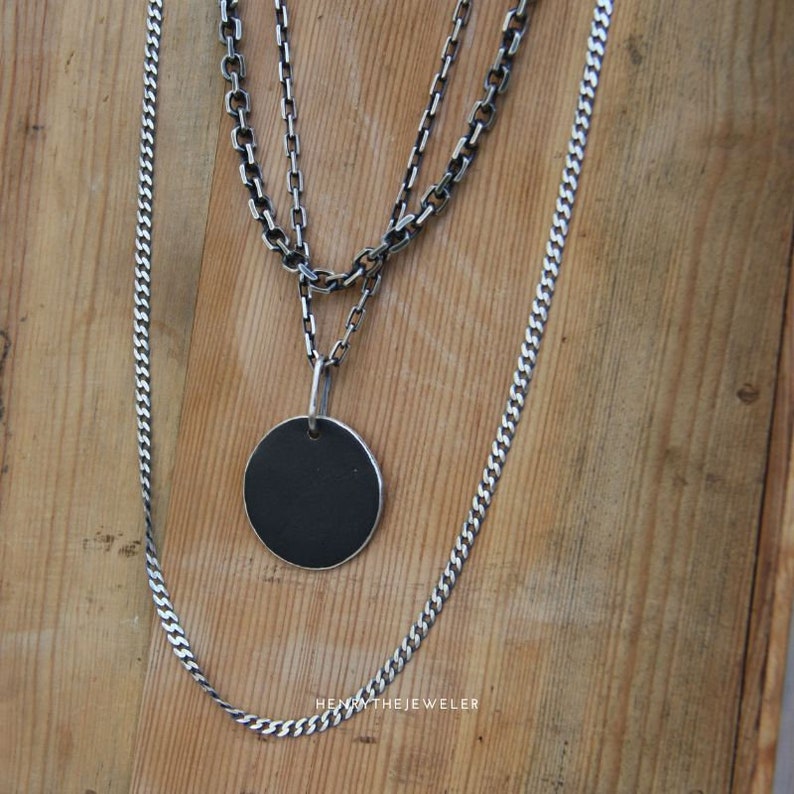 May include: Three silver chains with a round, black pendant. The chains are layered on top of each other and the pendant is hanging from the bottom chain. The chains are on a wooden surface.