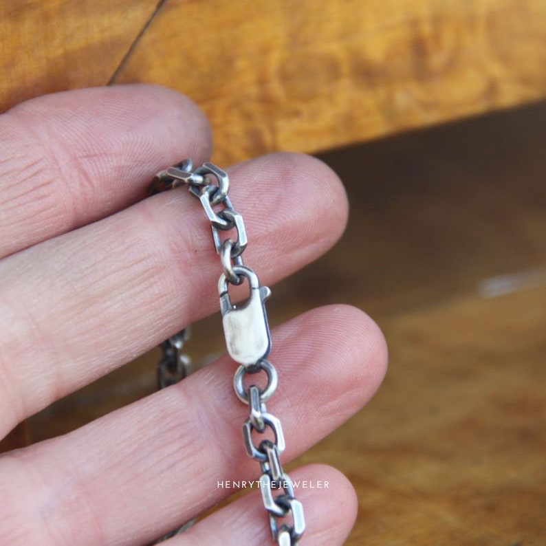 May include: A silver chain necklace with a lobster clasp closure. The chain is made of small, square-shaped links.