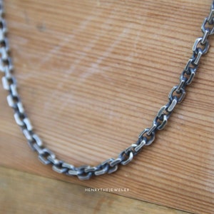 May include: A silver chain necklace with a box chain design. The chain is on a wooden surface.