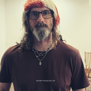 May include: A man with long, wavy hair and a beard is wearing a brown t-shirt, a silver skull necklace, and a red and white knit hat. He is also wearing glasses. The text "HENRYTHEJEWELER" is at the bottom of the image.