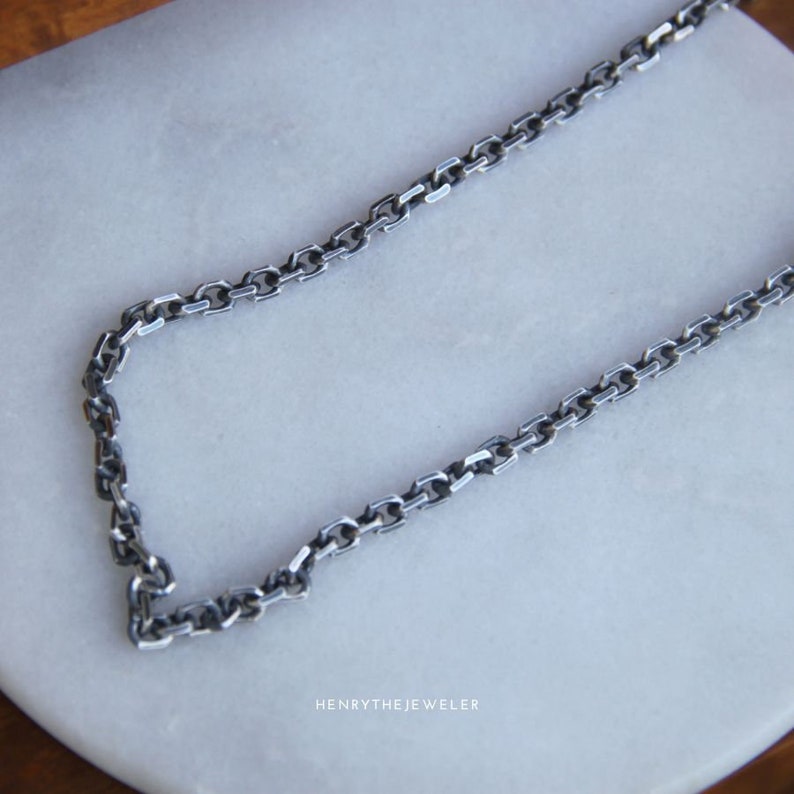May include: A silver chain necklace with a box chain design. The chain is on a white surface.
