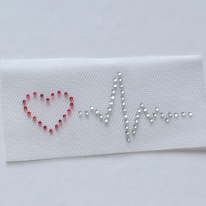 Könnte beinhalten: Ein weißer Stoff-Patch mit einem roten Herzen und einem silbernen Strass-Herzschlag-Design.