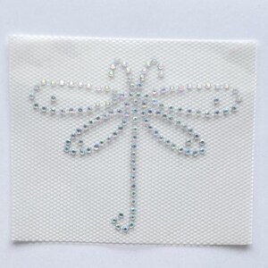 May include: A white square patch featuring a dragonfly design. The dragonfly is created with iridescent rhinestones, arranged to form the insect's body and wings. The patch has a textured, woven background.