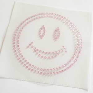 May include: A white fabric patch with a pink rhinestone smiley face design.