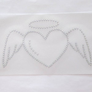 May include: A white fabric with a silver rhinestone design of an angel with wings, a halo, and a heart.