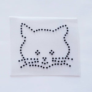 May include: A white iron-on transfer sheet with a black outline of a cat face. The cat has two black dots for eyes and a black line for a nose.