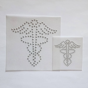Puede incluir: Dos parches de tela blanca con diseños de caduceo médico en strass plateados. El parche más grande mide aproximadamente 10 cm de ancho y el más pequeño aproximadamente 5 cm de ancho.