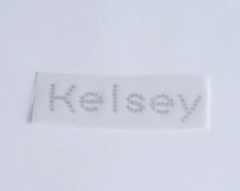 May include: A white fabric patch with the name "Kelsey" spelled out in silver rhinestones.