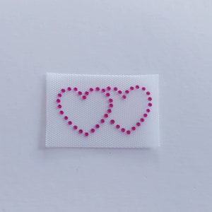 Op de afbeelding: Twee roze strass harten op een witte stoffen achtergrond.