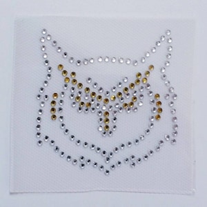 Op de afbeelding: Een vierkant wit stoffen embleem met een uilontwerp van strass-steentjes. De omtrek van de uil wordt gevormd door zilveren strass-steentjes, met gouden strass-steentjes die het midden van het ontwerp accentueren. De strass-steentjes vormen het gezicht en het lichaam van de uil.