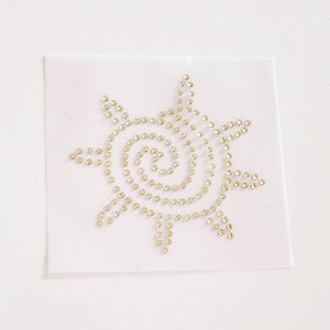 May include: A white fabric patch with a gold rhinestone sun design. The sun has a spiral pattern in the center.