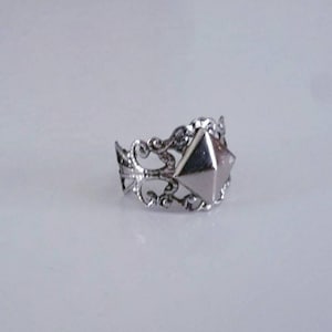 May include: A silver-toned ring with an ornate, filigree band and a central, geometric, pyramid-shaped design. The ring's design features swirling patterns and a pointed, multi-faceted center stone, creating a unique and eye-catching accessory.