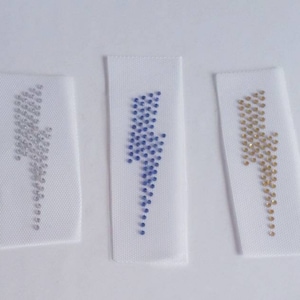 Może przedstawiać: Trzy białe łatki z tkaniny z wzorem błyskawicy wykonanym z cyrkonii. Błyskawice są srebrne, niebieskie i złote.