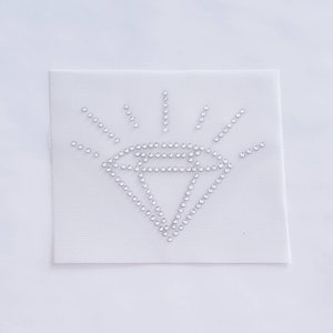 May include: A white fabric patch with a diamond shape made of silver rhinestones. The diamond has a starburst design around it.