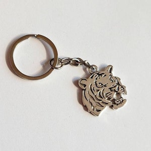 May include: Silver key chain with a detailed tiger head charm. The charm is facing to the right and has a detailed design of the tiger's face.