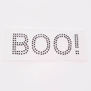 May include: A white background with the word "BOO!" spelled out in black rhinestones.