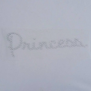 May include: The word "Princess" spelled out using small, clear rhinestones on a white background.
