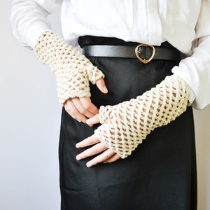 May include: A pair of off-white crocheted fingerless gloves with a delicate open-weave pattern. The gloves are worn by a person wearing a white button-down shirt and a black skirt with a heart-shaped belt buckle.