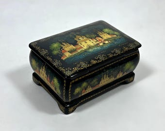 May include: A black lacquered box with a hinged lid, featuring a hand-painted landscape scene of a village with yellow buildings, green trees, and a blue river. The box has gold decorative trim and rests on four small feet.