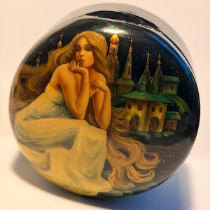 May include: A round black lacquer box featuring a hand-painted scene of a person with long flowing blonde hair wearing a white dress, sitting beside a dark green building with multiple spires and crosses against a dark starry night sky.