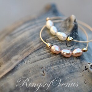May include: Two gold bracelets with pearl accents. The top bracelet has white pearls and the bottom bracelet has pink pearls. The bracelets are on a textured, gray surface. The text "Rings of Venus" is visible in the bottom right corner of the image.