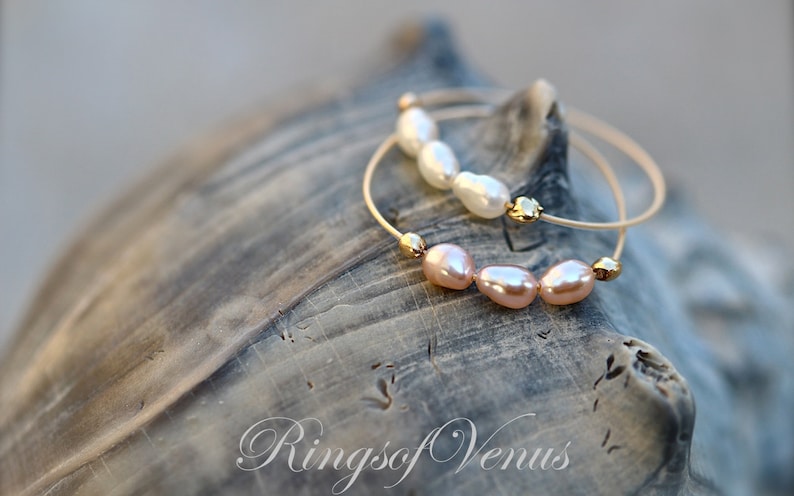 May include: Two gold bracelets with pearl accents. The top bracelet has white pearls and the bottom bracelet has pink pearls. The bracelets are on a textured, gray surface. The text "Rings of Venus" is visible in the bottom right corner of the image.